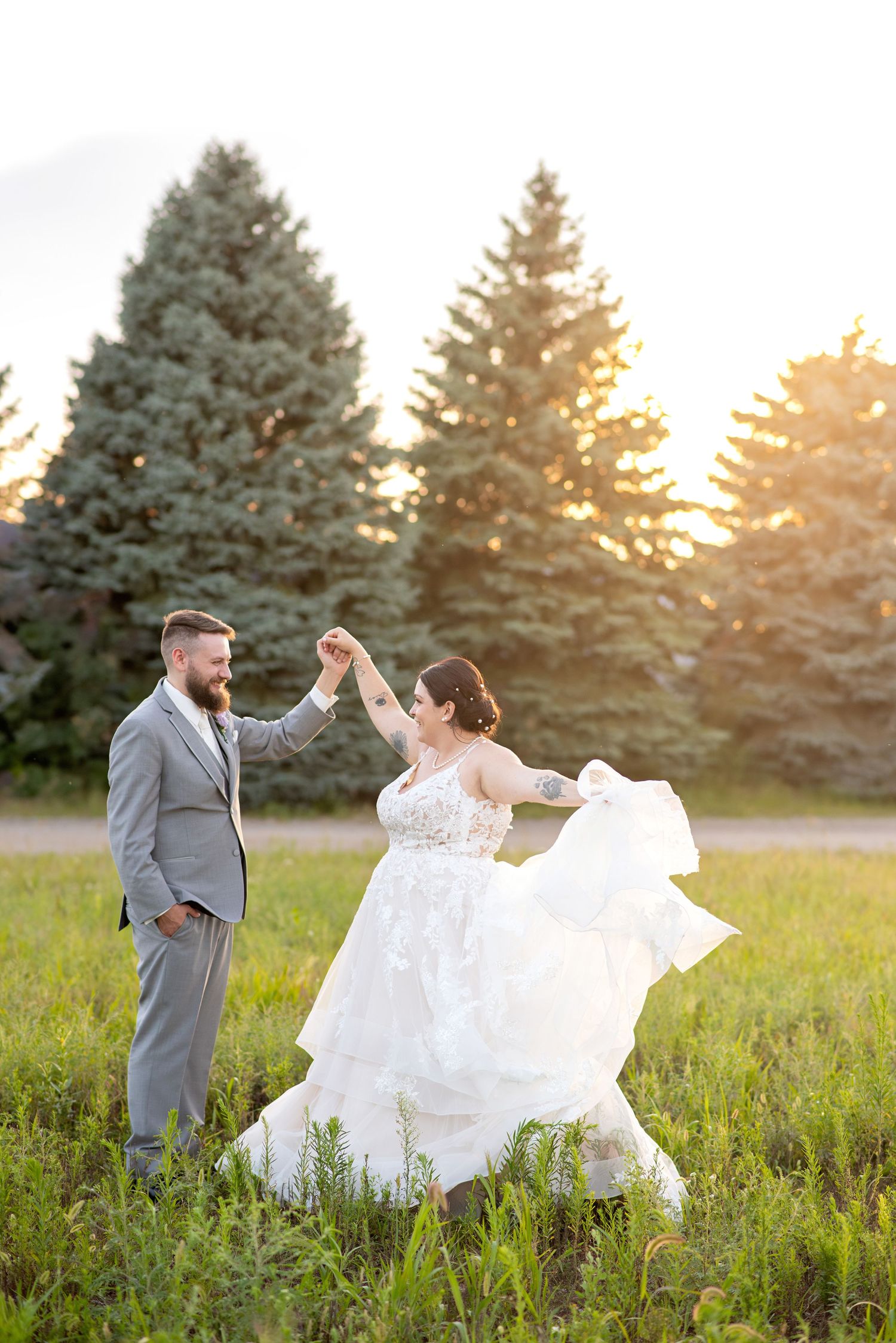 Bride and groom dancing in a sunlit field with evergreen trees, bride's gown flowing in the breeze.