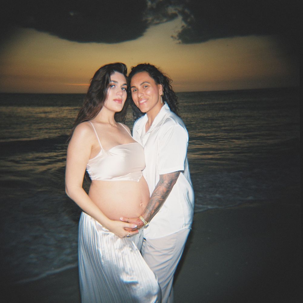 Couple posing for maternity photo on beach at sunset, woman in white dress showing baby bump.