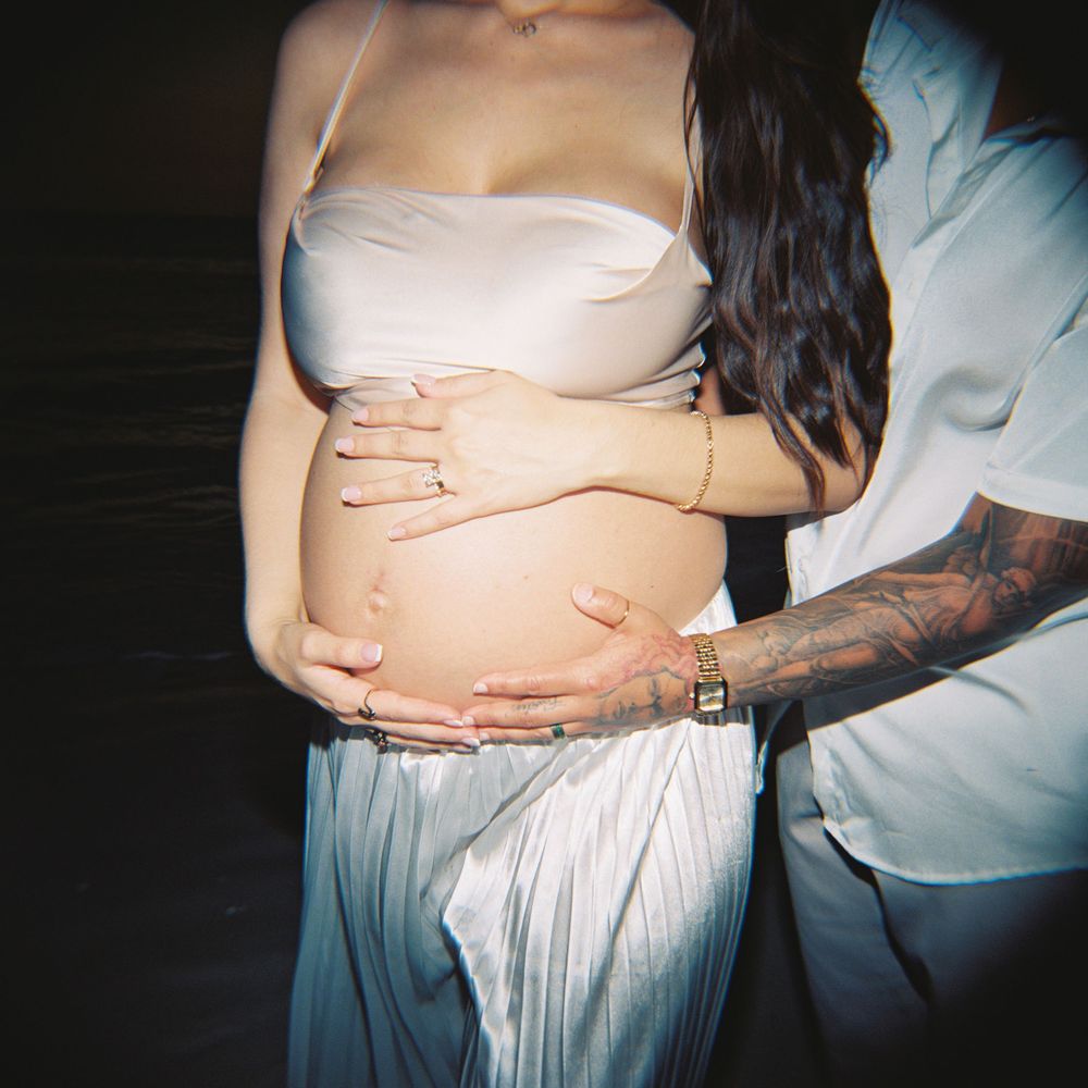 Close-up of a pregnant woman's baby bump with tattooed partner's hands gently cradling her belly.