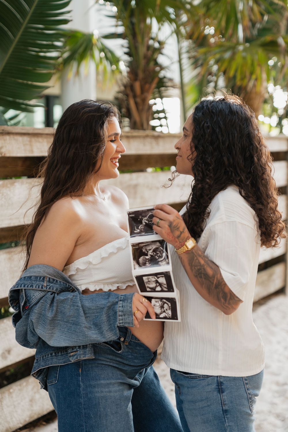 Two women smile at each other while holding ultrasound photos outdoors near palm trees.