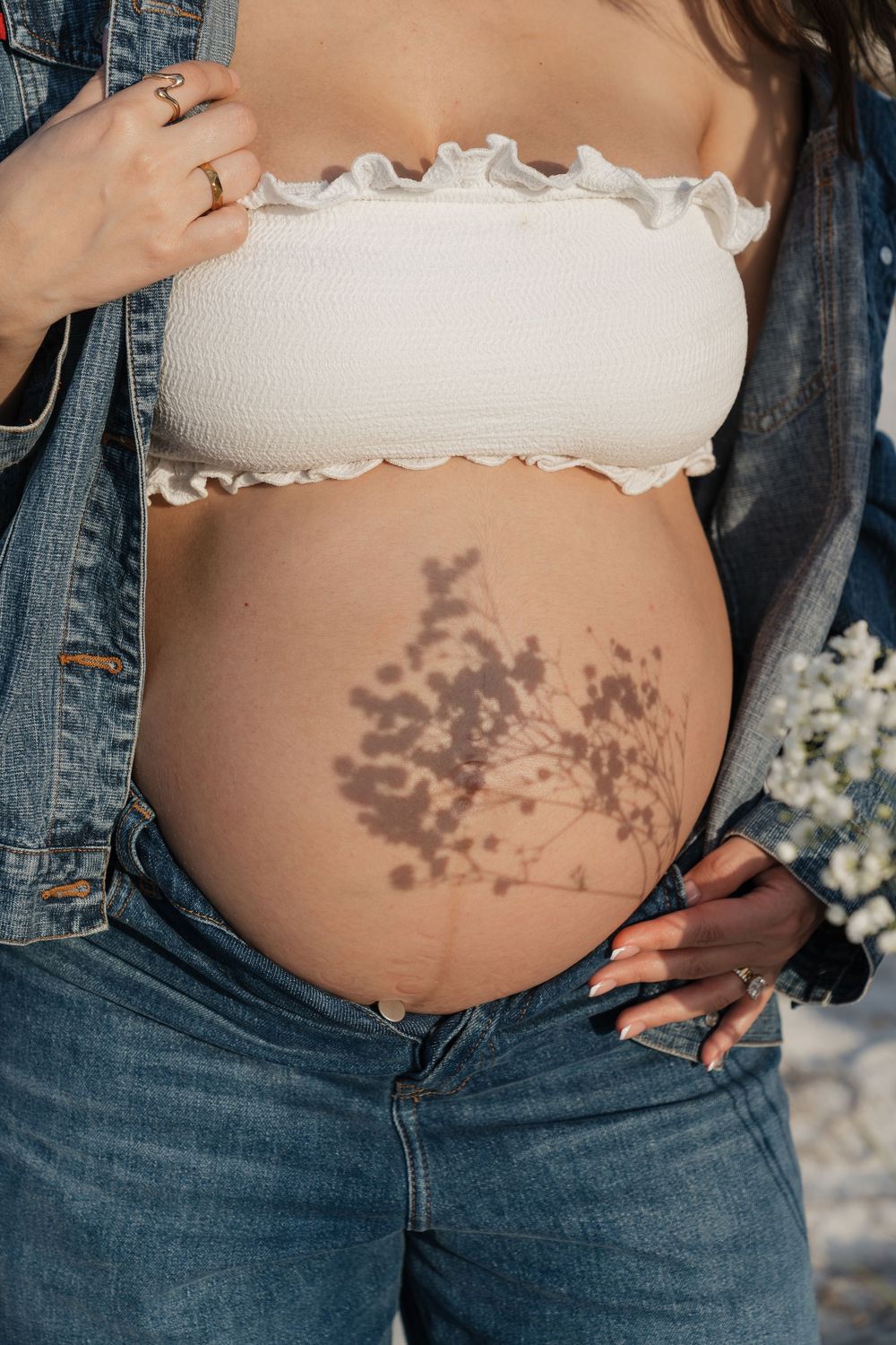 Pregnant woman in white bandeau top and denim jacket with floral shadow on baby bump.