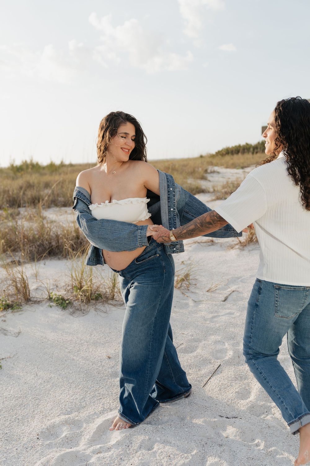 Two women playfully interact on white sand beach, one pregnant in denim jacket and jeans, the other in white top.