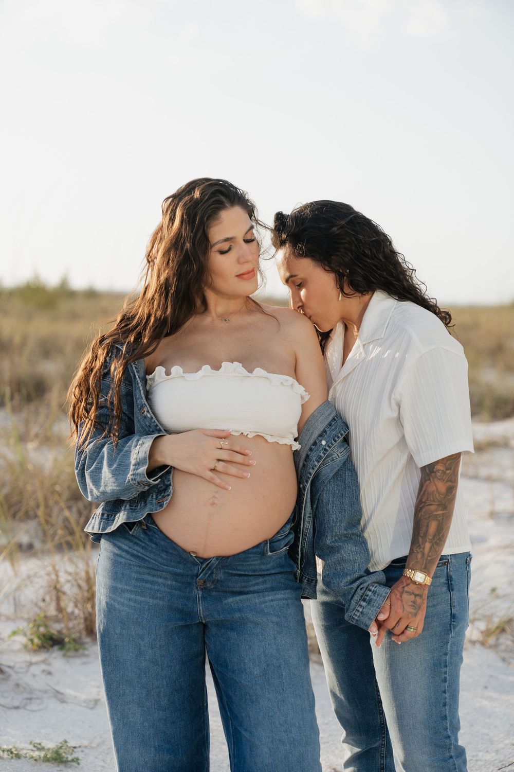 Pregnant woman in white tube top and jeans shares tender beach moment with partner kissing her baby bump.