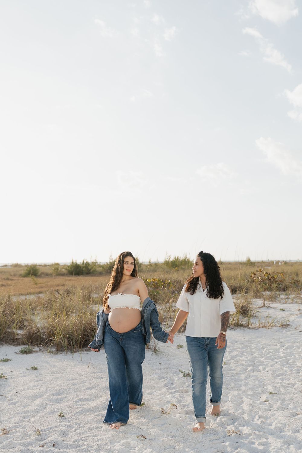 Pregnant woman in denim jeans holding her baby bump while walking on white sand beach with her partner.