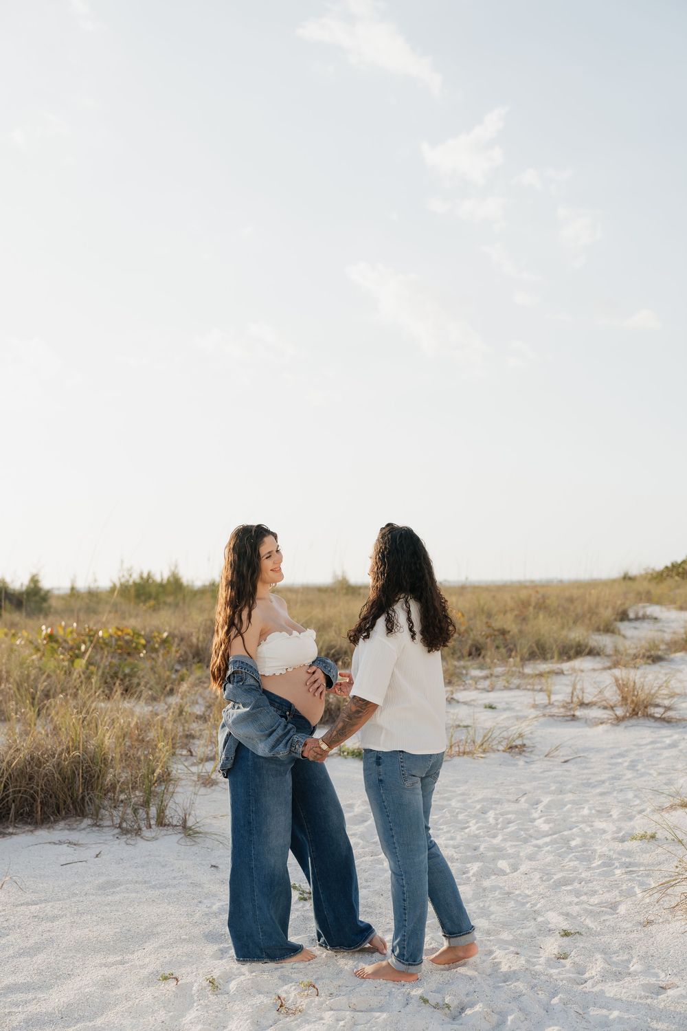 Two women stand together on sandy beach, one visibly pregnant in denim jacket, both wearing jeans at sunset.