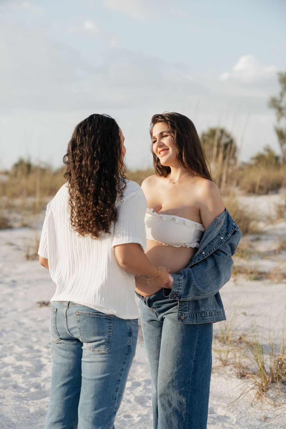 Two women in jeans share a tender moment on a white sand beach, one pregnant, both smiling softly.