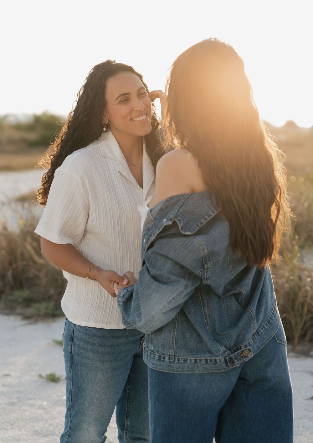 Two women share an intimate moment on a sunlit beach, one smiling in white shirt while other faces away.