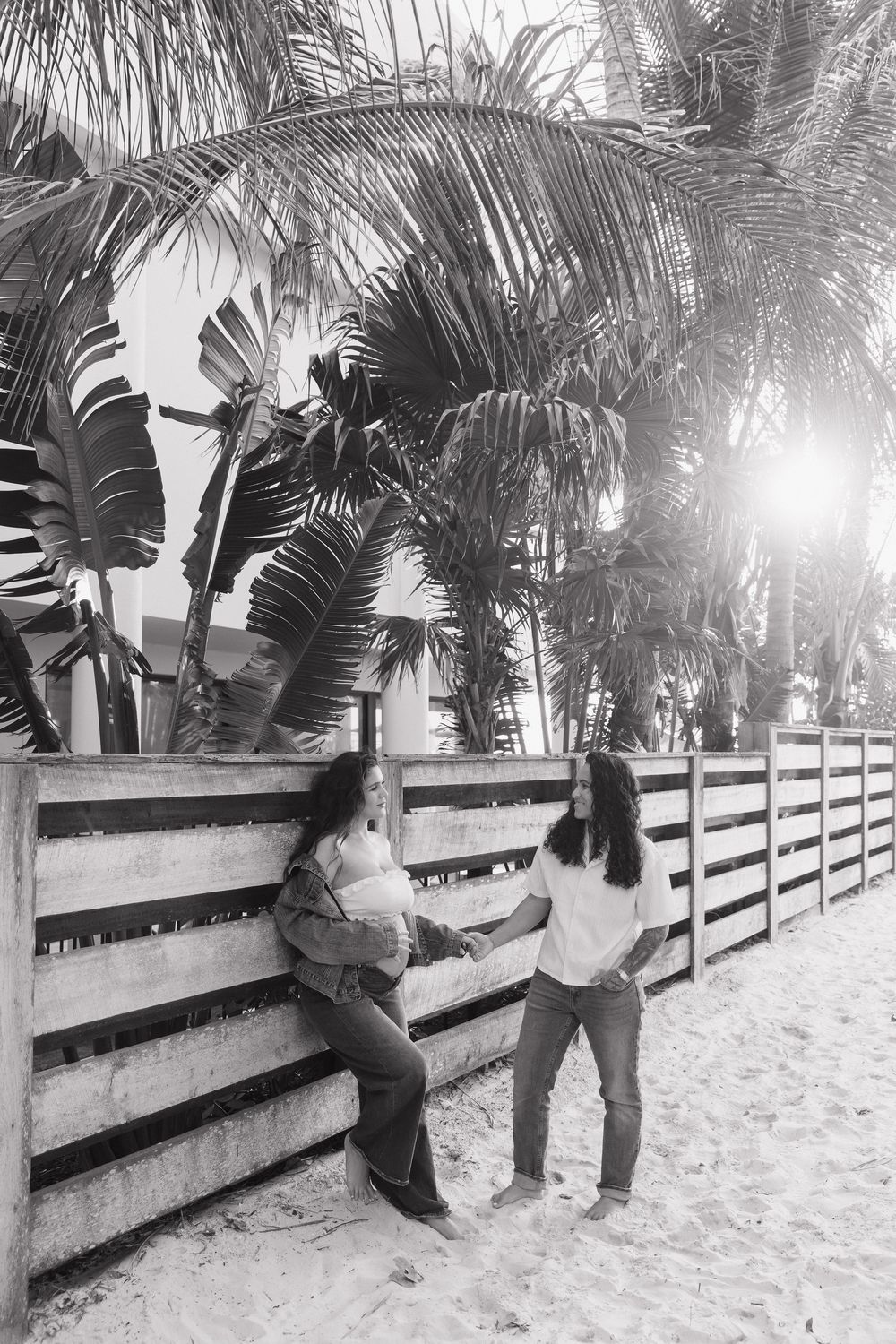 Black and white photo of two women talking by a wooden fence on a sandy beach with tropical palm trees.