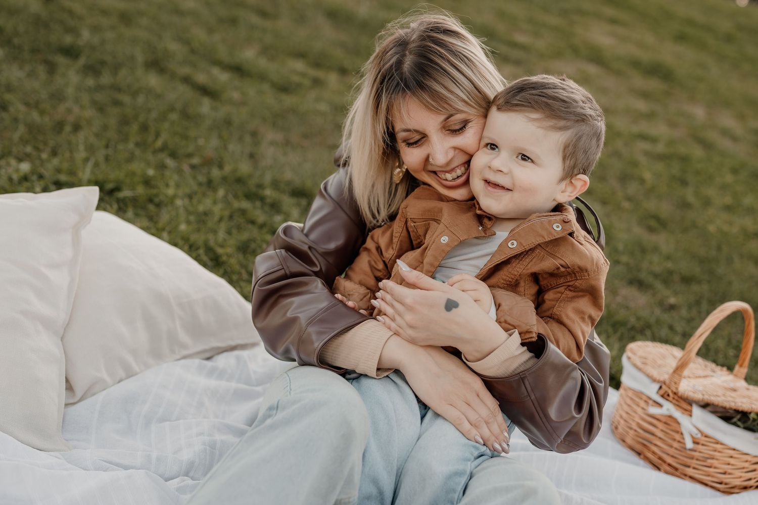 A smiling mother hugs her happy toddler son on a white picnic blanket in a green grassy park.