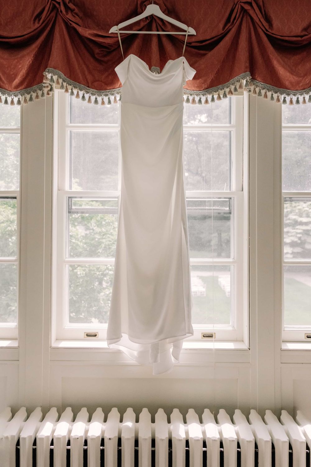 Elegant white satin wedding dress hanging in a window with red curtain valance.