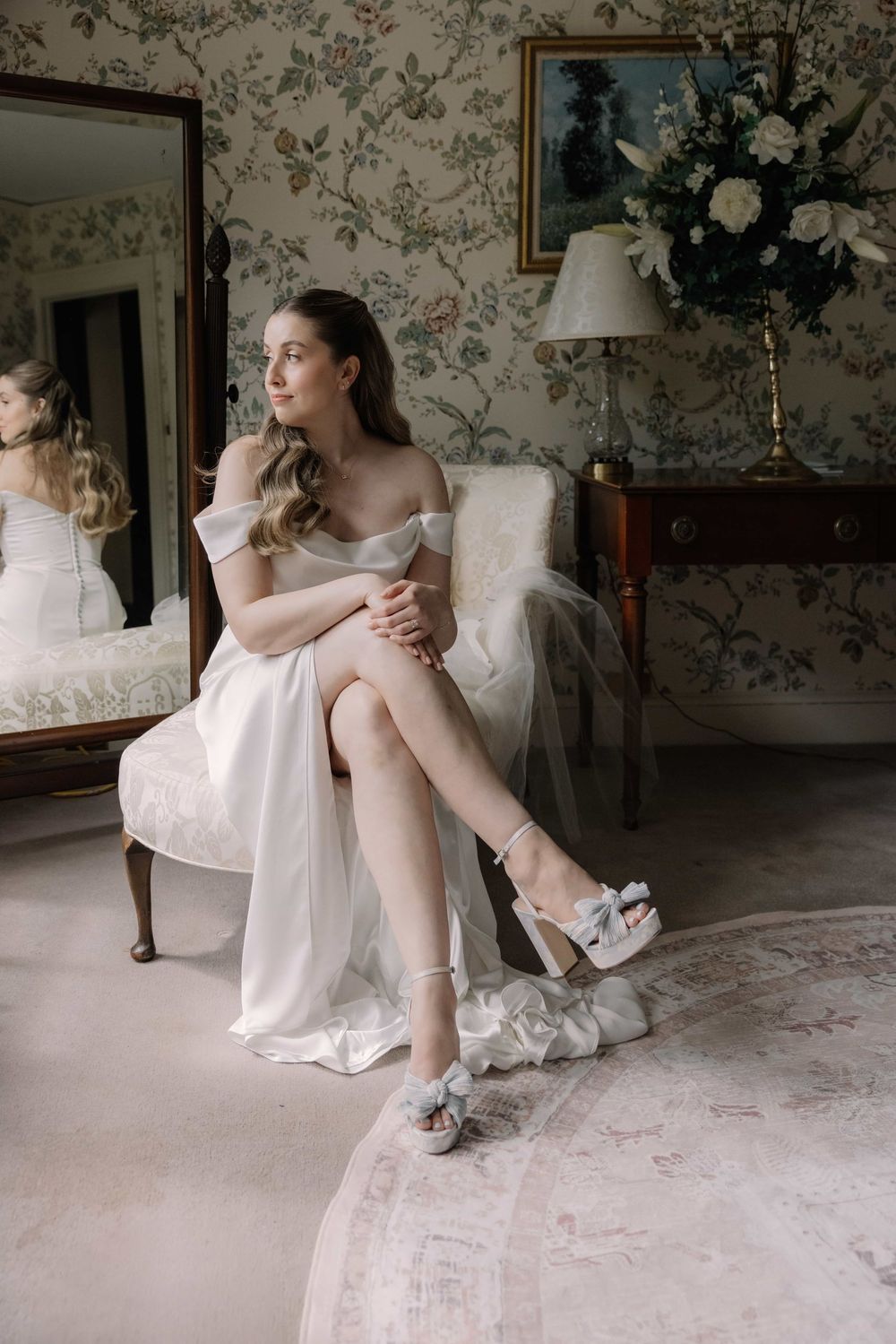 Elegant bride in off-shoulder white gown seated in vintage armchair wearing bow heeled sandals in floral wallpapered room.