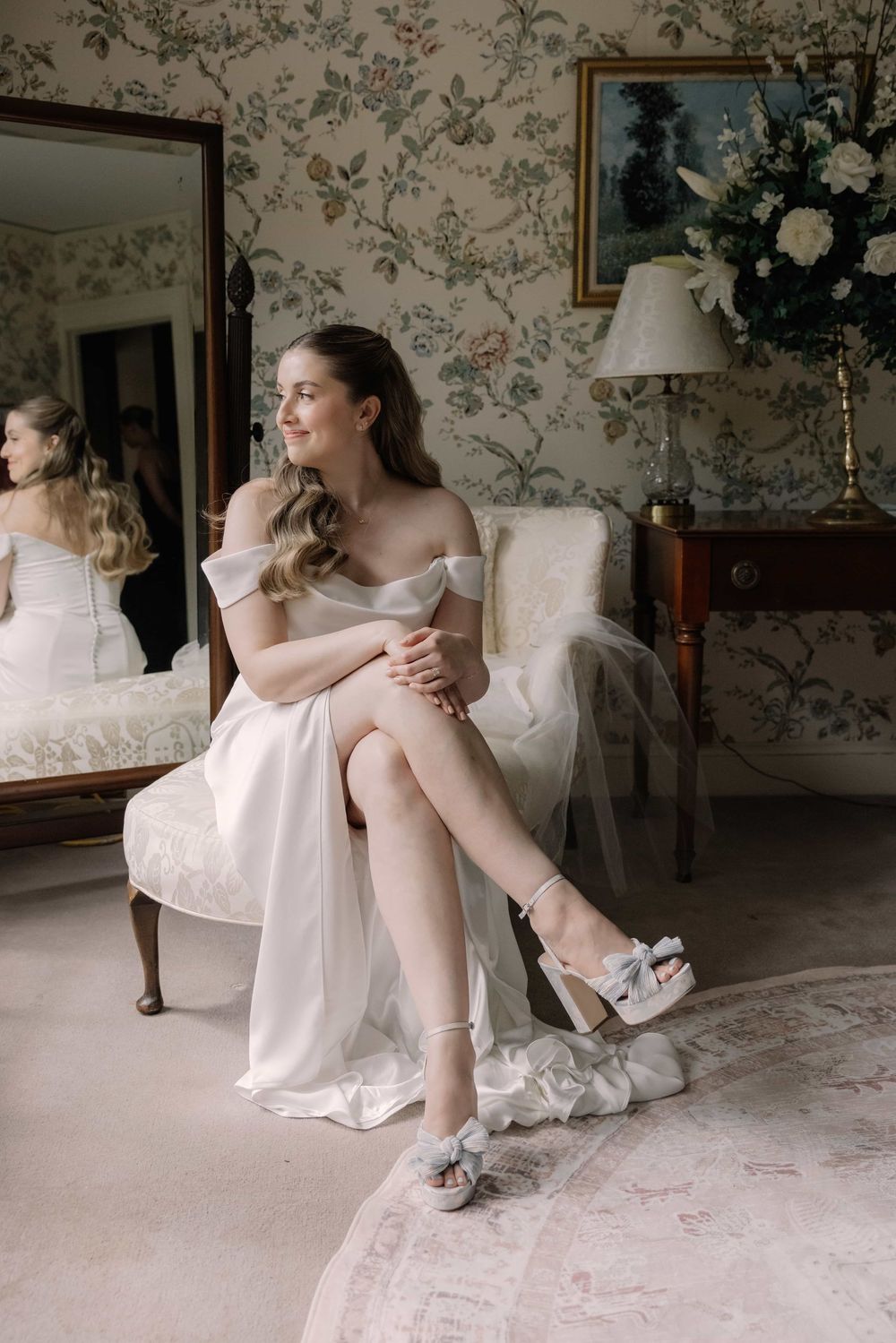 Radiant bride in off-shoulder white dress seated in armchair wearing bow heeled sandals in elegant floral bridal suite.