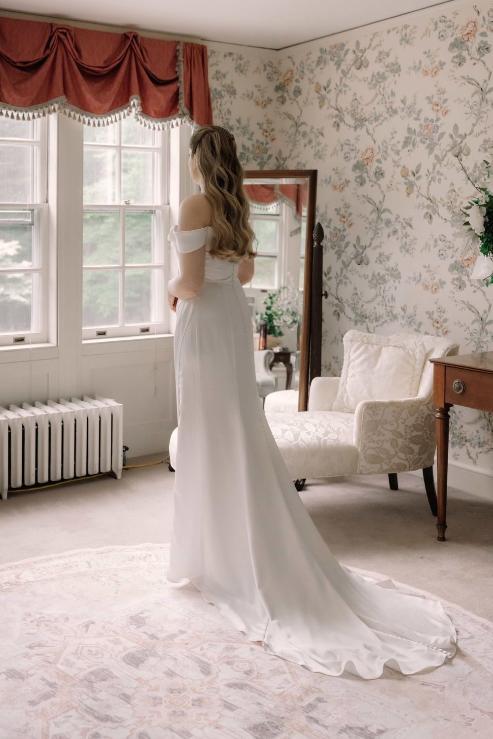Bride in off-shoulder white wedding gown with train standing by window in vintage floral wallpapered bridal suite.