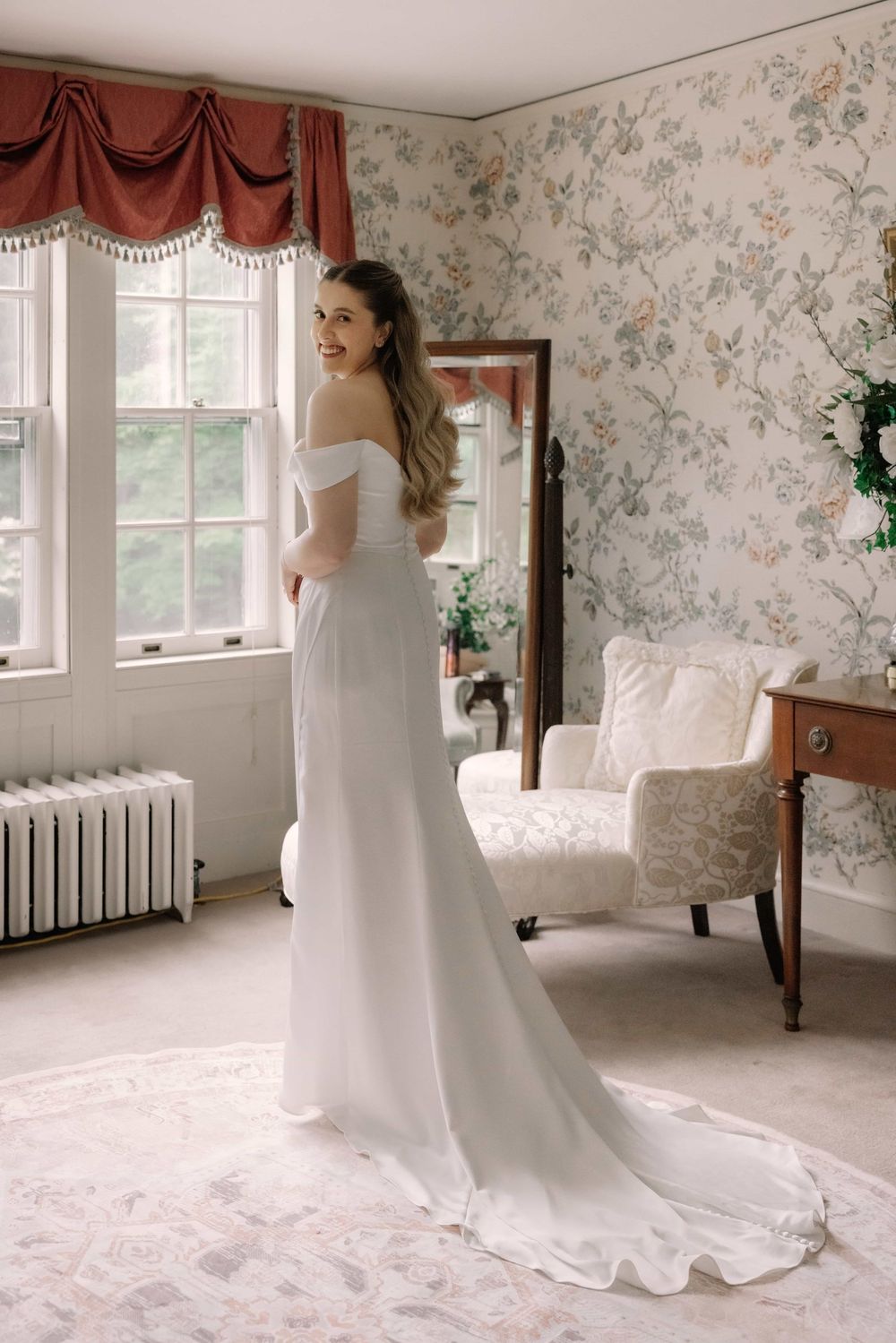 Bride in elegant off-shoulder white gown with train smiling over shoulder in vintage floral wallpapered bridal suite.