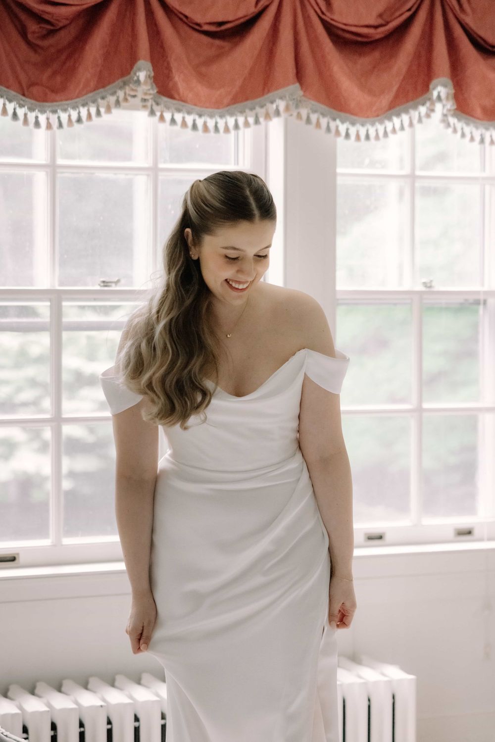 Joyful bride in off-shoulder white wedding gown smiling softly in front of large sunlit window.
