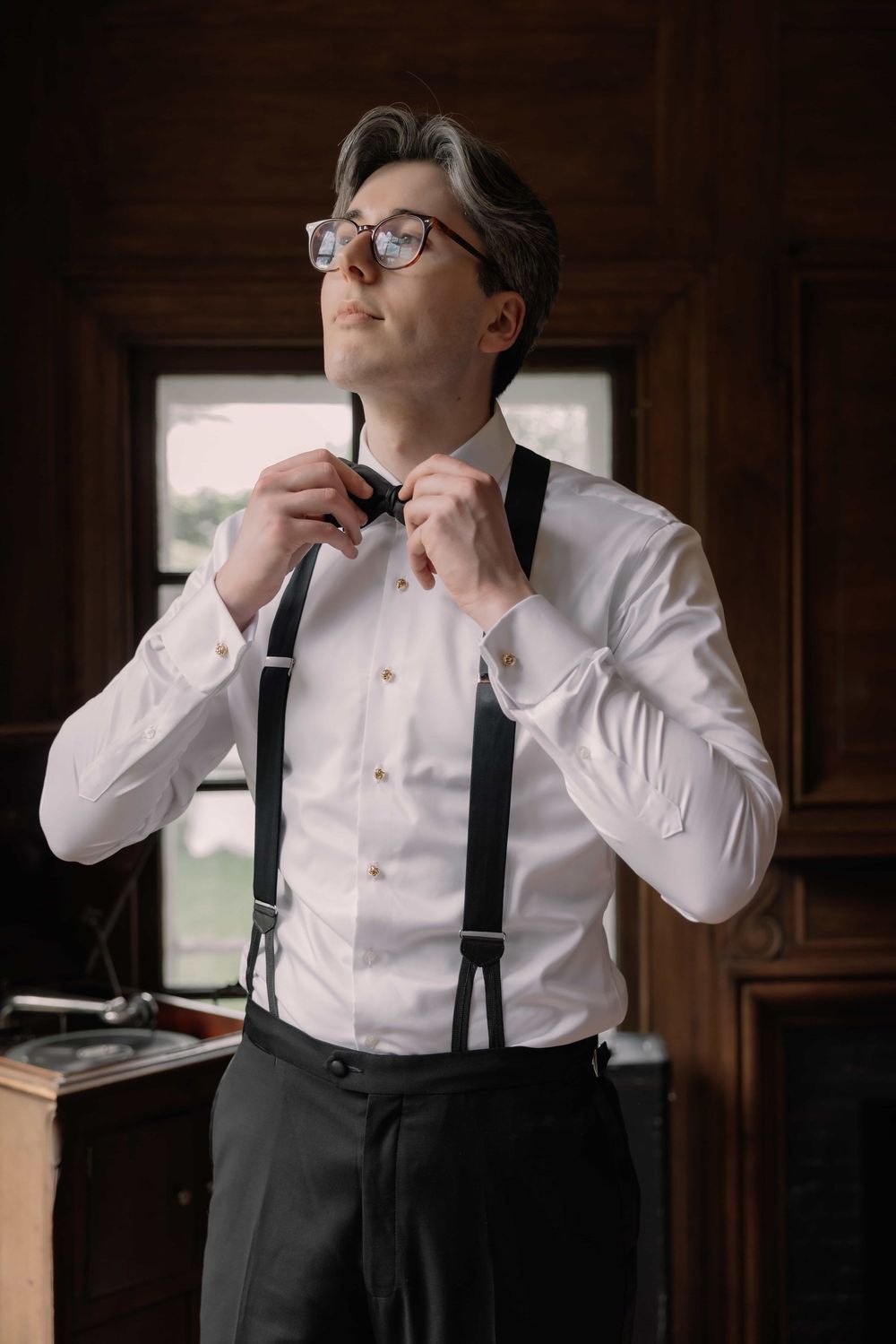 Groom in white shirt and suspenders adjusting bow tie while getting ready in a wood-paneled room.