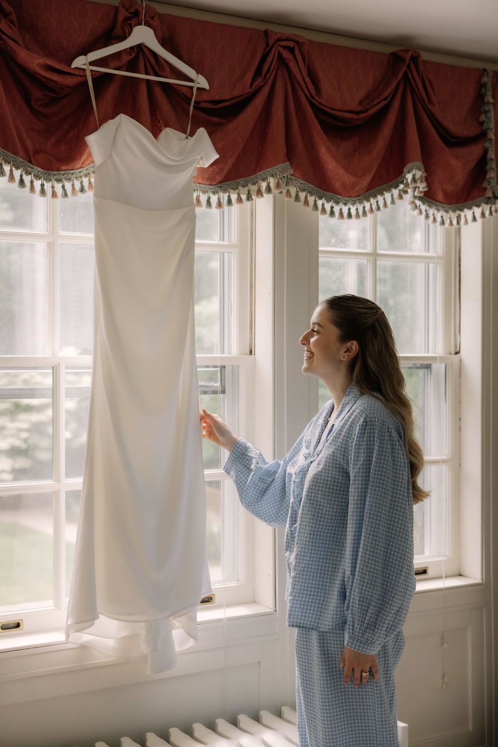 Bride in blue pajamas smiling at her white wedding dress hanging in a sunlit window.