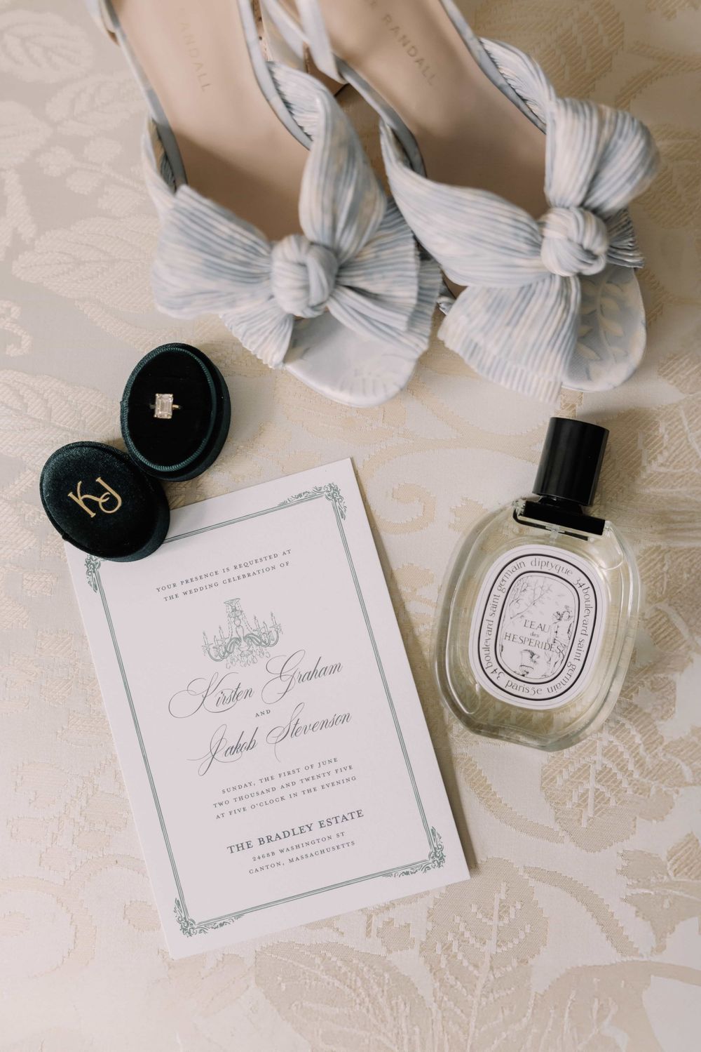 Bridal flat lay featuring bow heeled sandals, velvet ring box, Diptyque perfume, and elegant wedding invitation on cream fabric.