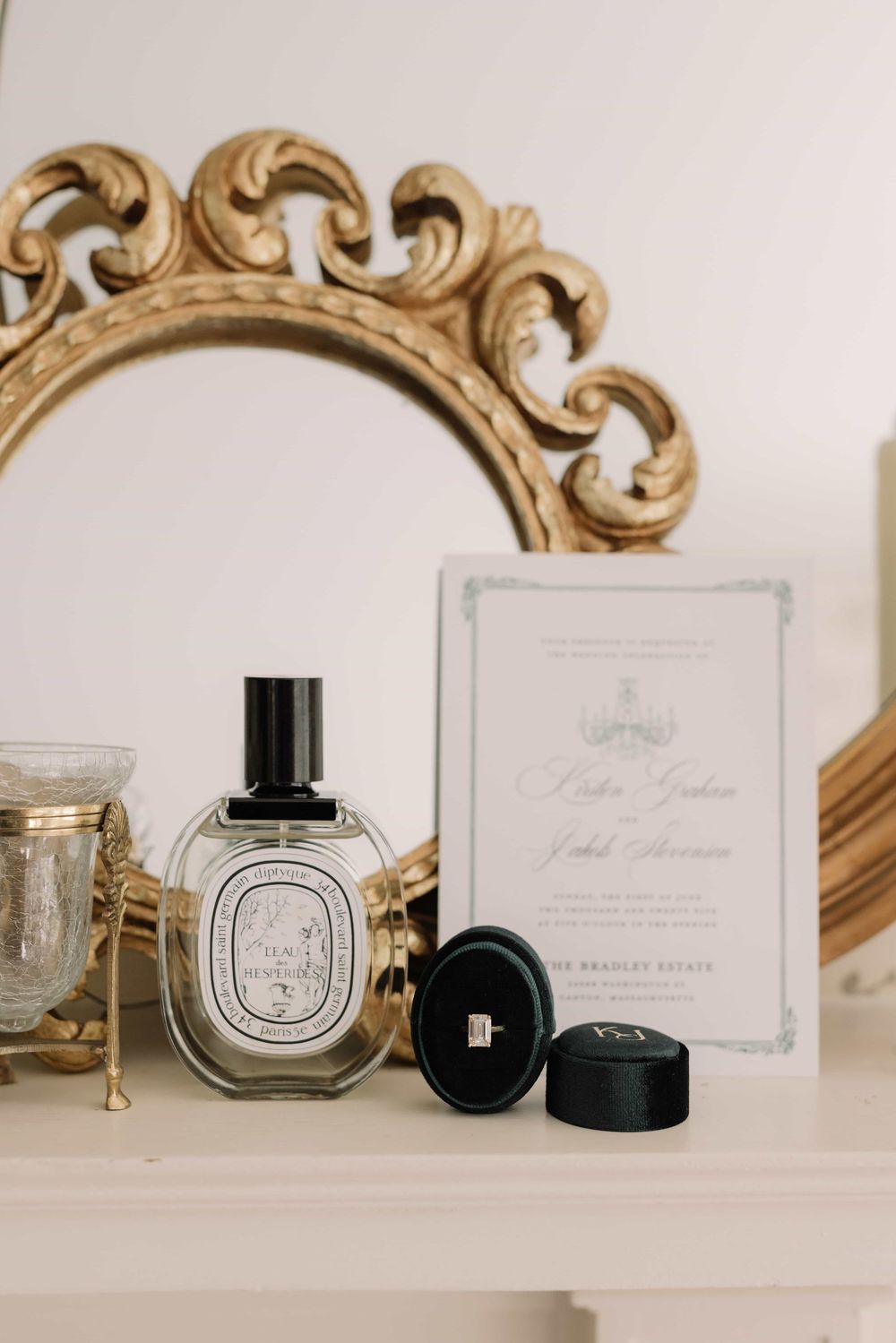 Elegant bridal flat lay with Diptyque perfume, diamond ring in velvet box, and wedding invitation near gold ornate mirror.