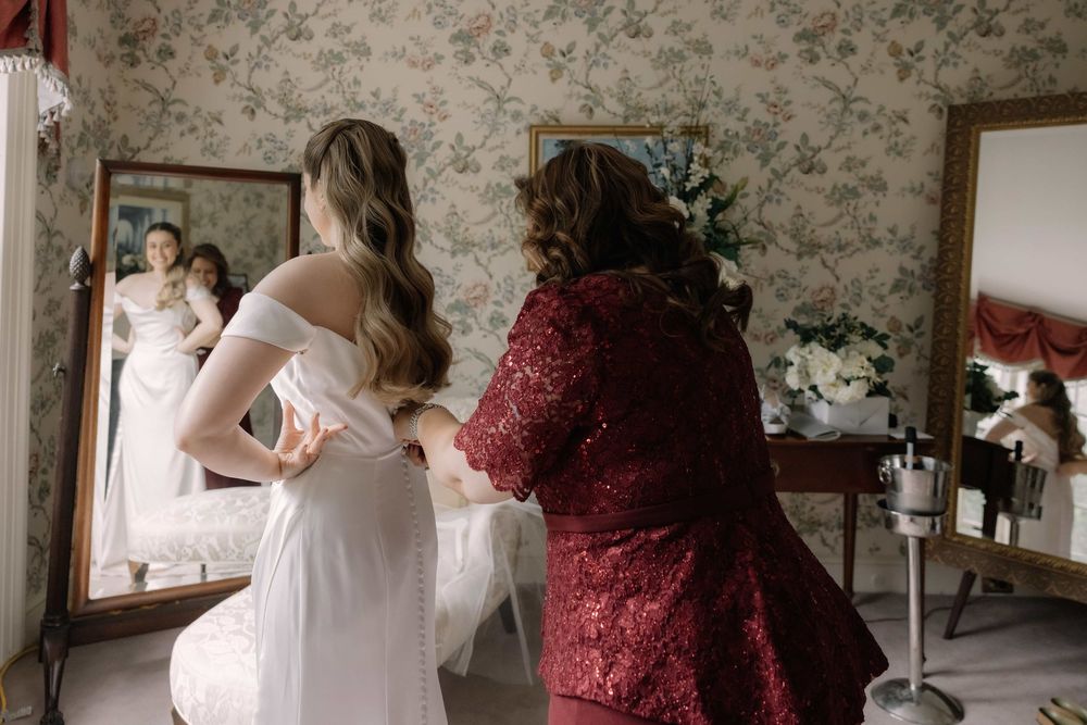 Bride in off-shoulder white gown being helped by woman in red lace dress in floral wallpapered dressing room.