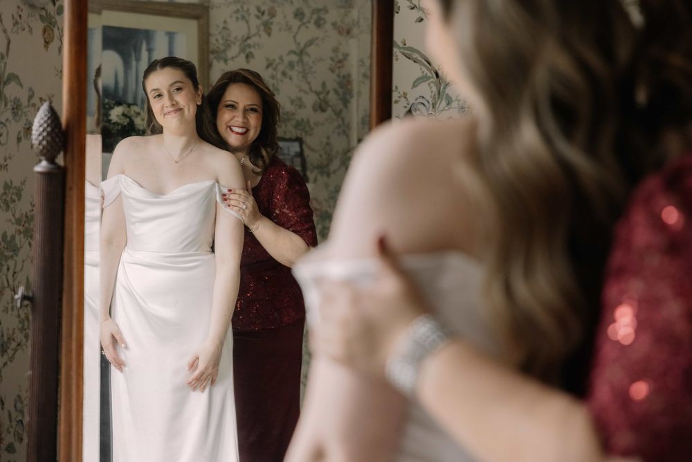 Smiling bride in off-shoulder white wedding gown with bridesmaid in red sequin dress reflected in vintage mirror.