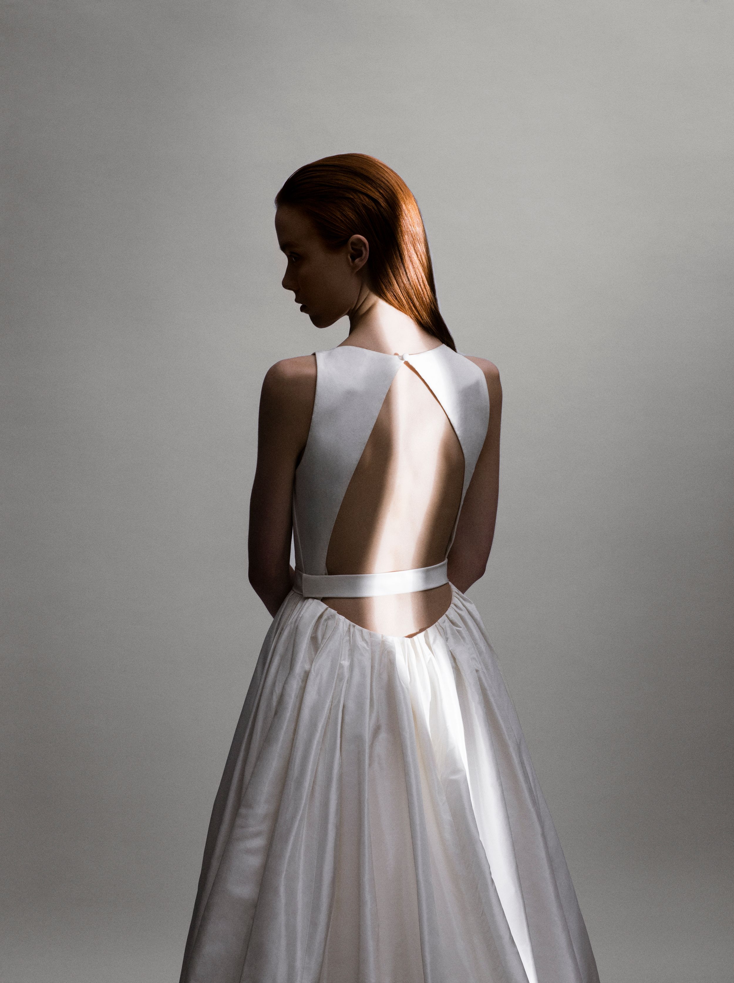 Elegant backless white wedding gown with geometric cutout detail, worn by a redhead model against a gray background.
