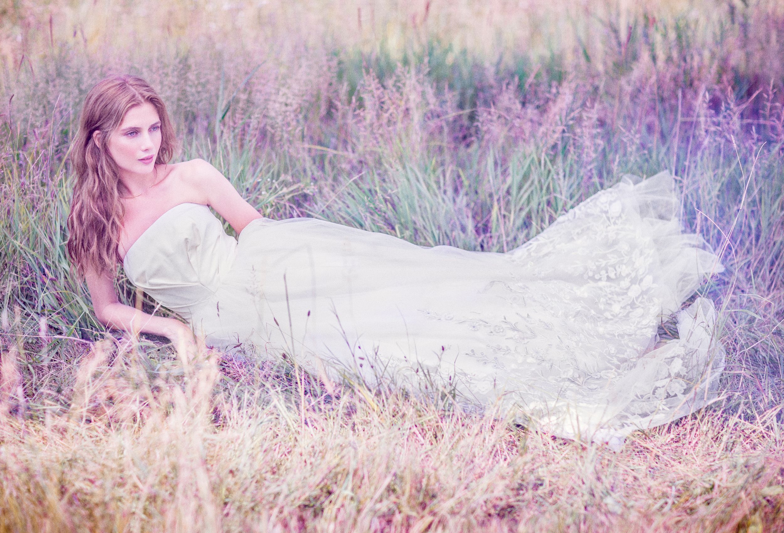 A woman in a flowing white lace gown reclines gracefully in a dreamy field of lavender wildflowers.