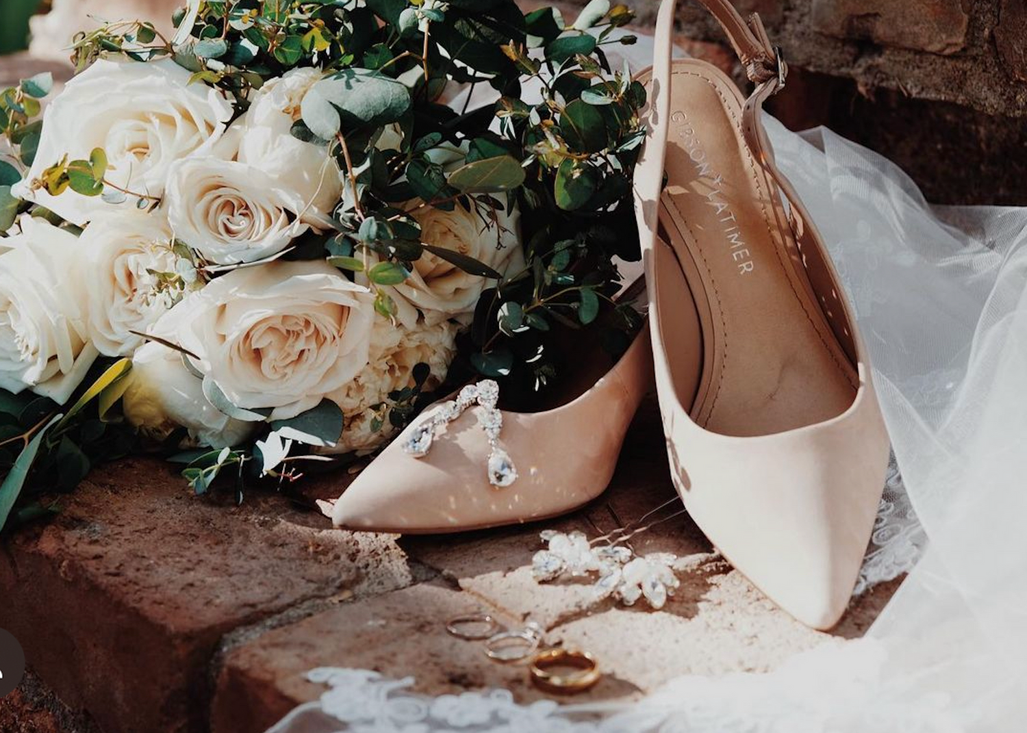 Wedding shoes in blush pink satin sit beside cream roses and greenery on rustic wooden steps.