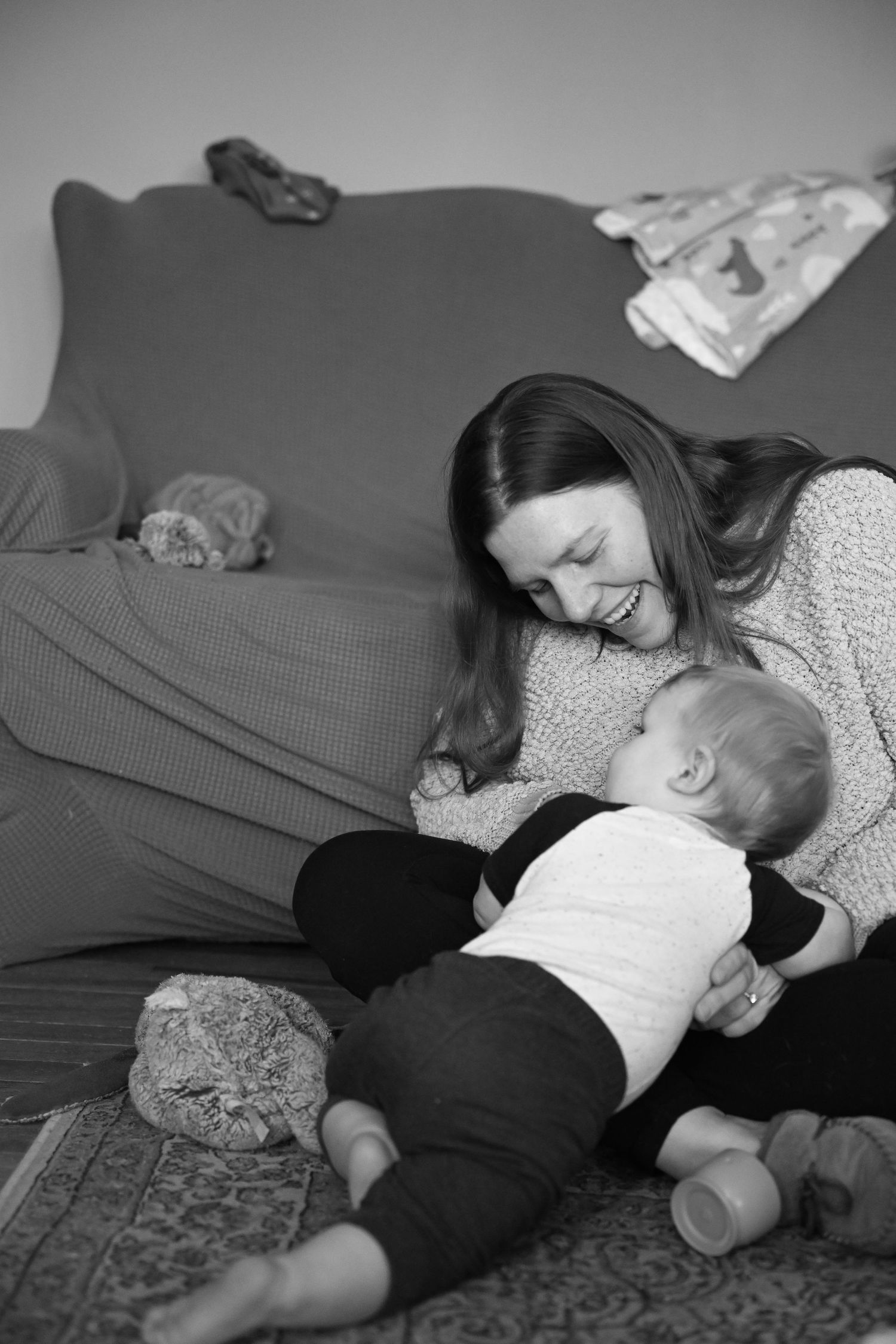 A black and white photo of Amber holding Nolan in the living room, sitting on the floor as he crawls around on her