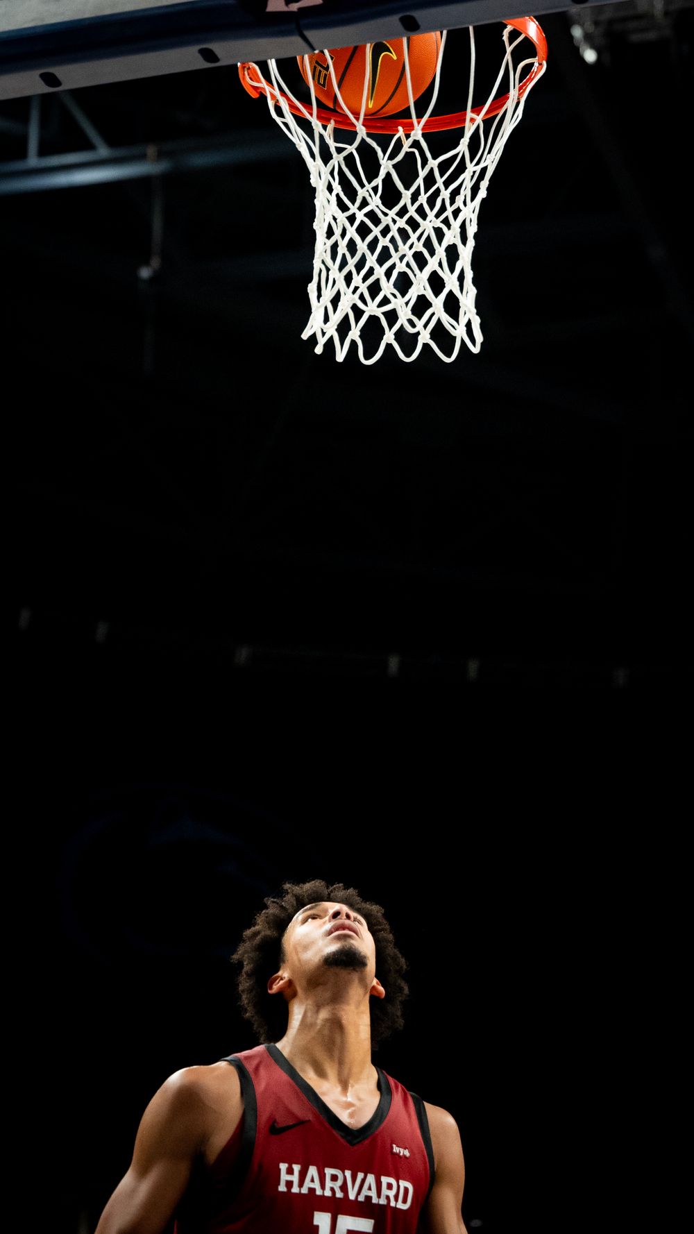 Basketball player in Harvard jersey looks up at the hoop during a game against a dark background.