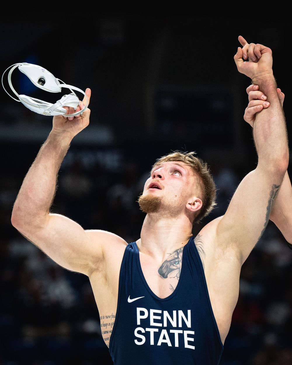 Penn State wrestler celebrates victory, raising hands and holding headgear in a navy blue Nike singlet.