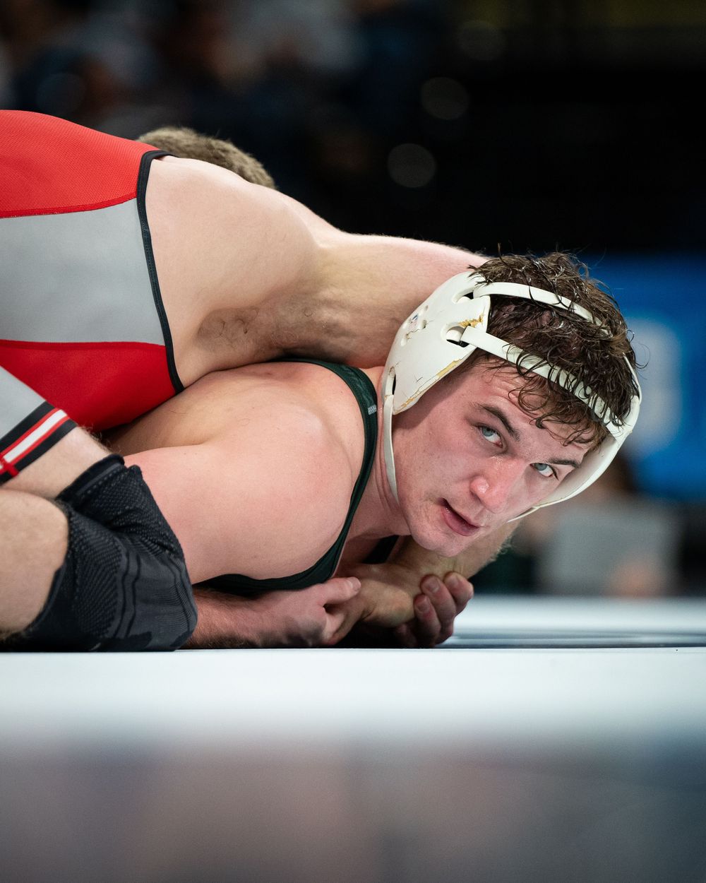 Two wrestlers compete in a match, with one athlete in a white headgear gaining control over his opponent on the mat.