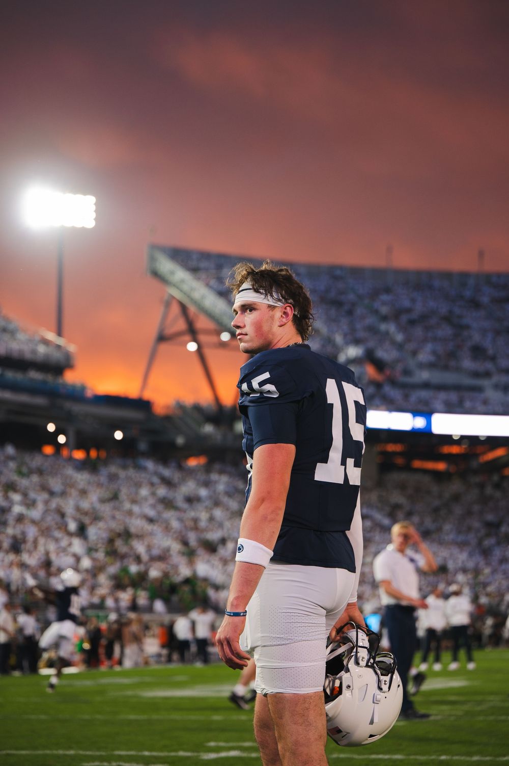 Football player stands on field at sunset wearing black jersey and white pants during pre-game warmups.