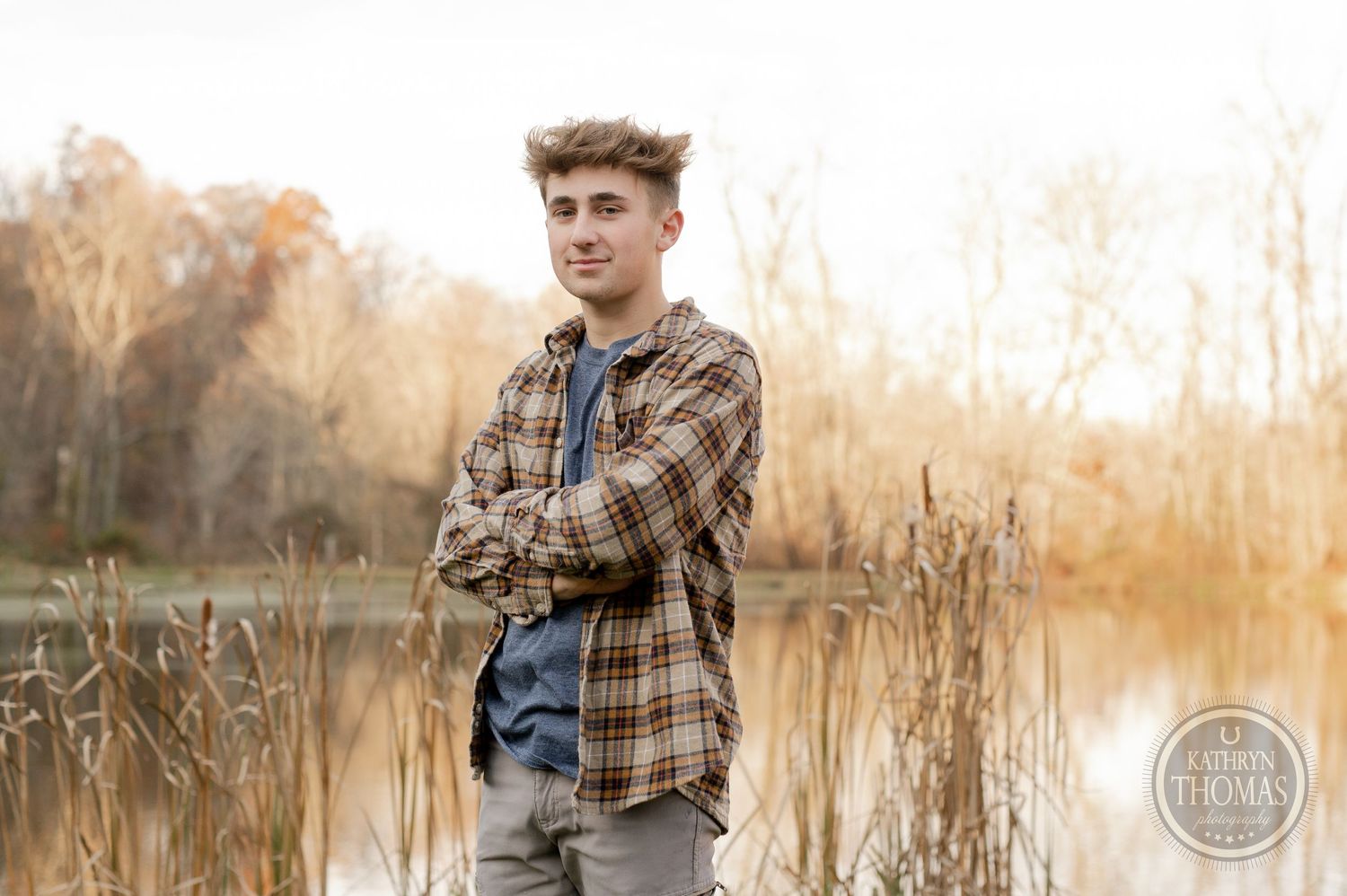 A person in a plaid flannel shirt poses with arms crossed by a lake during golden hour.