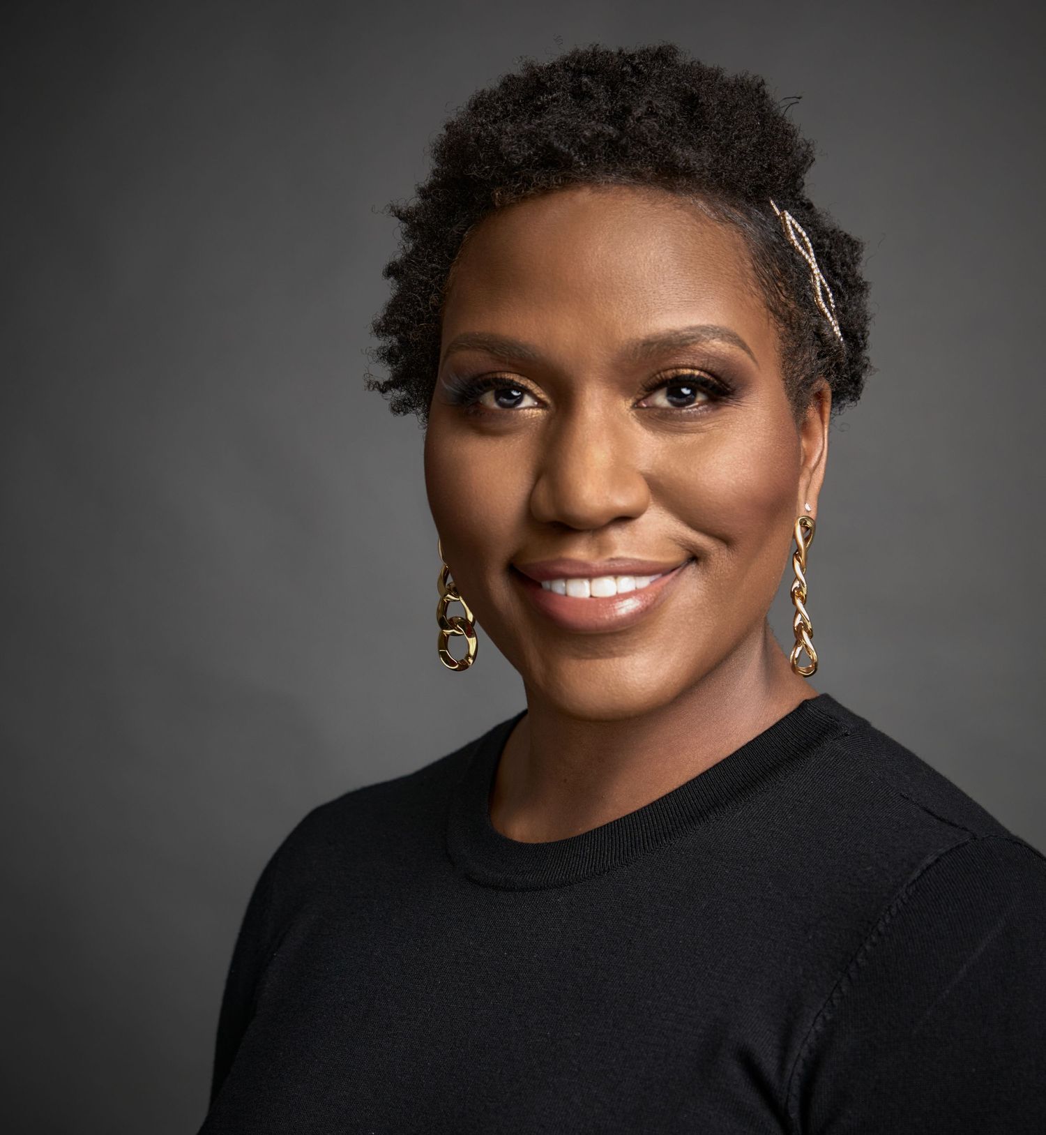 Studio portrait in black sweater with statement earrings against dark background.