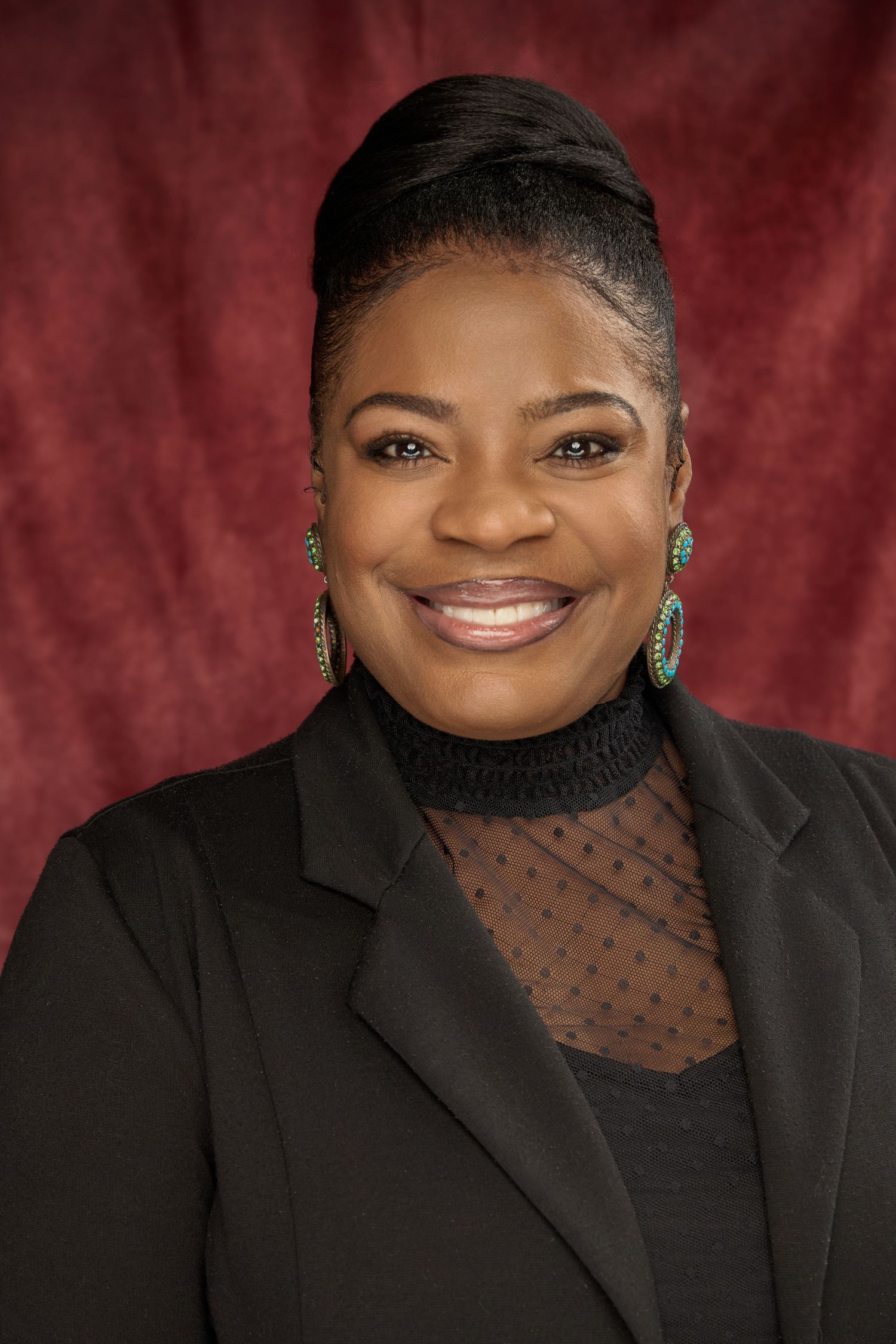 Professional portrait in black outfit with sheer details against deep red backdrop.