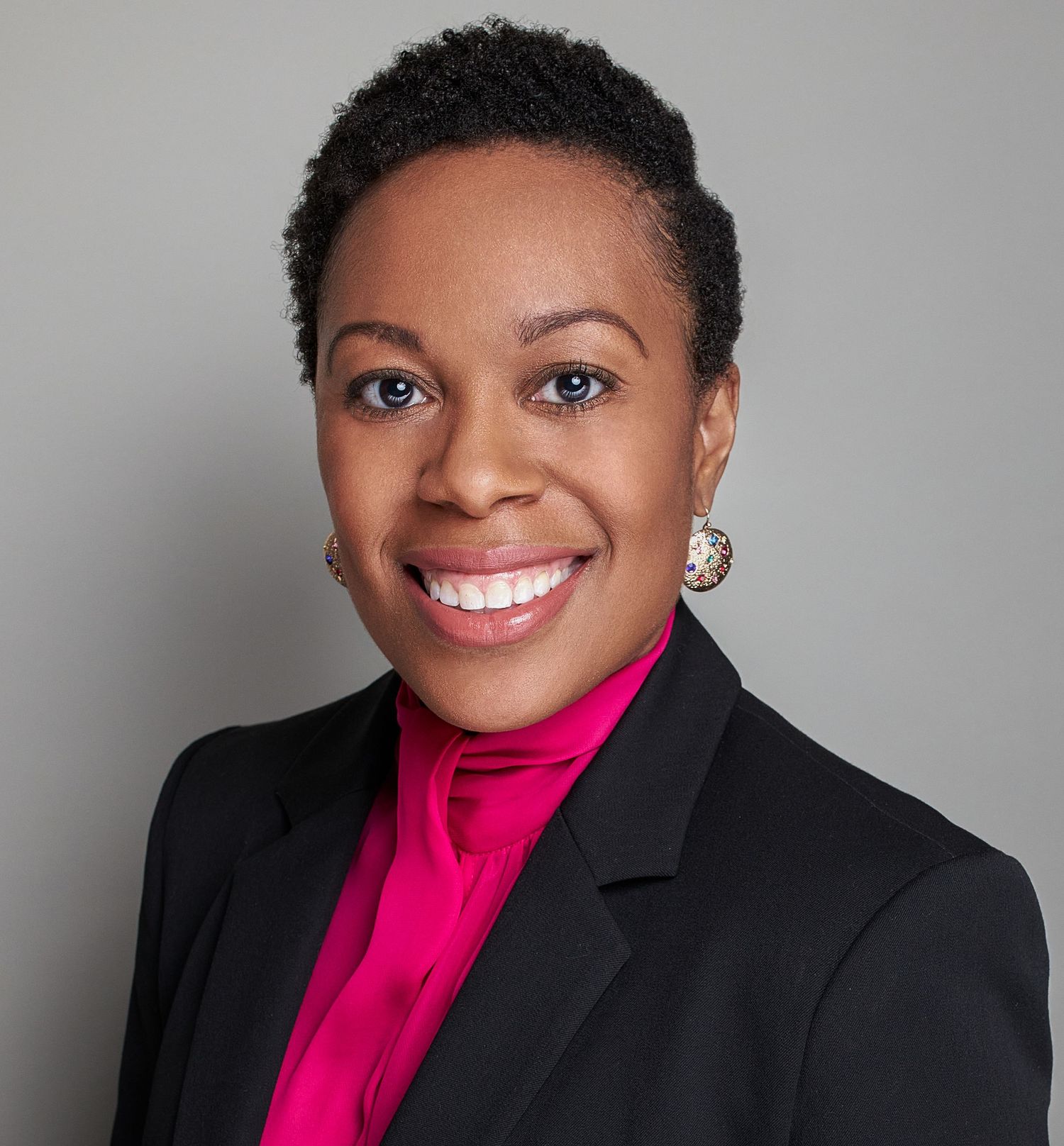 Professional portrait in black blazer and bright pink top against grey background.