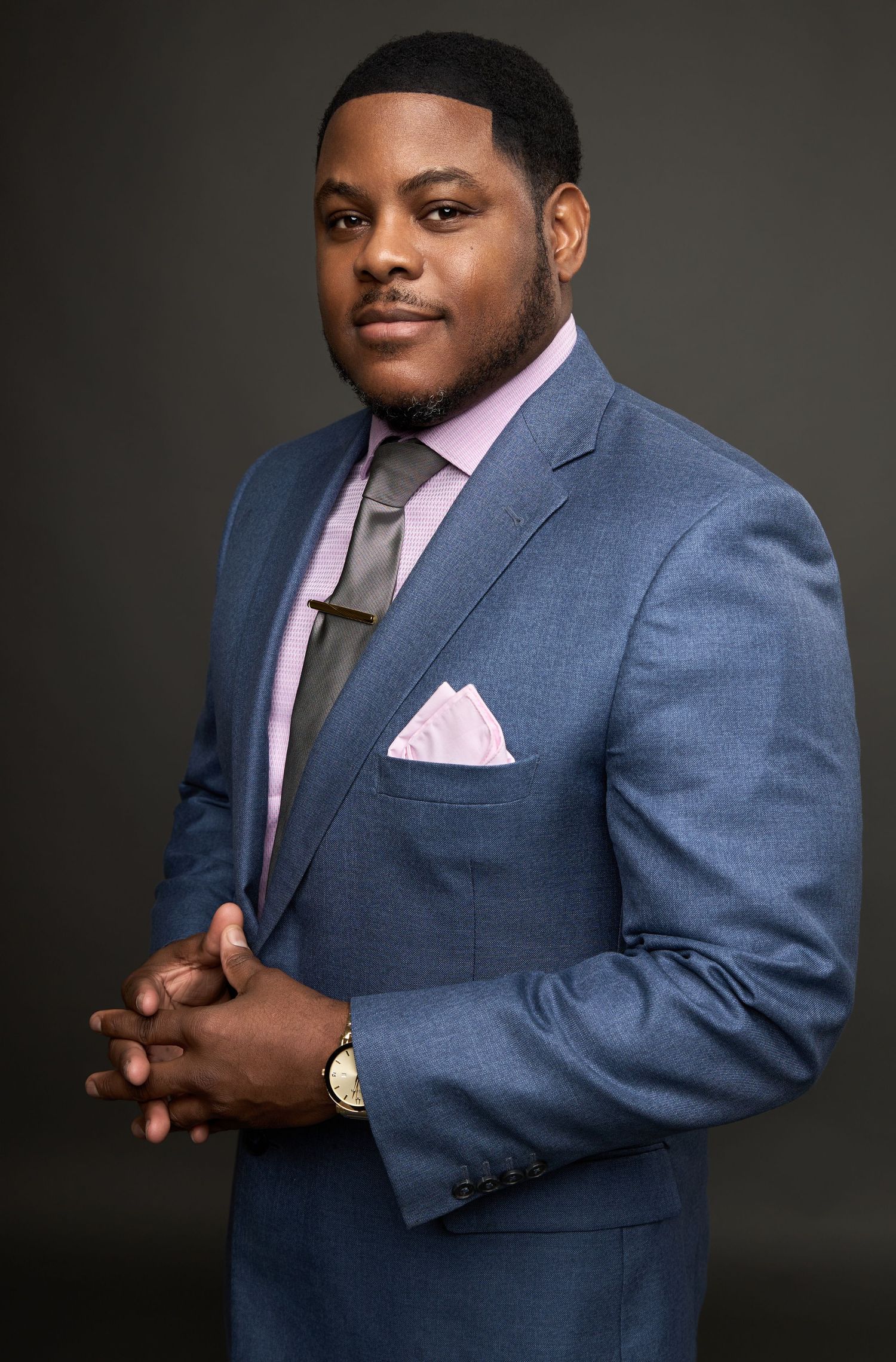 Professional portrait in blue suit with pink tie and pocket square against dark background.