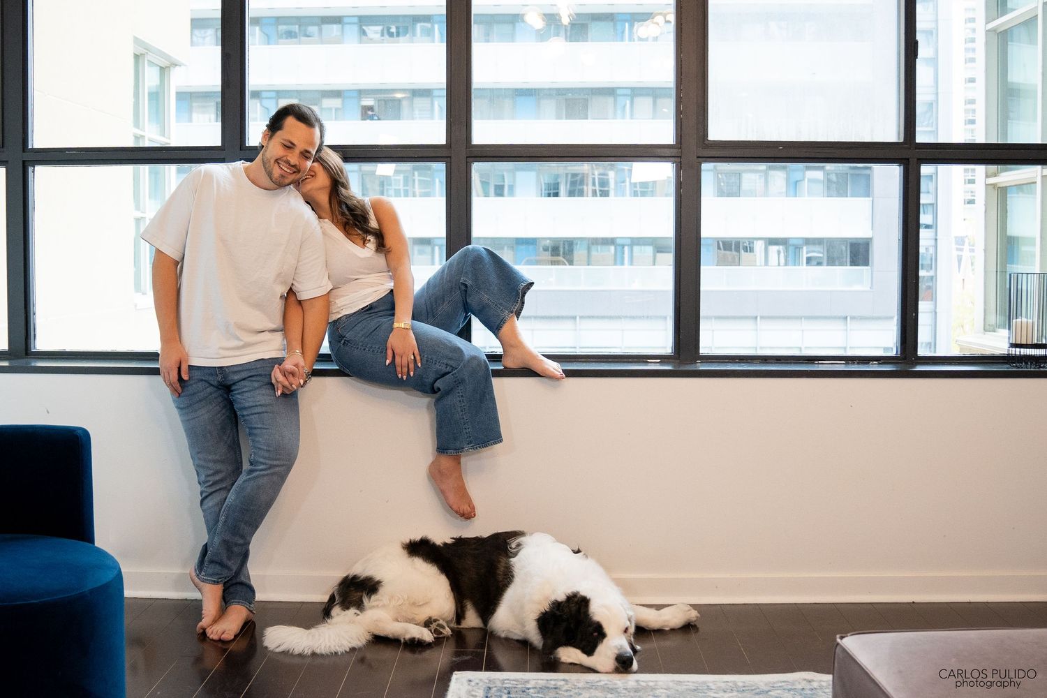 A couple lounging by a window with their black and white dog in a modern apartment setting.