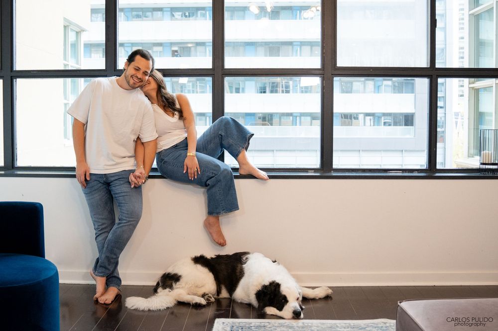 A couple lounging by a window with their black and white dog in a modern apartment setting.
