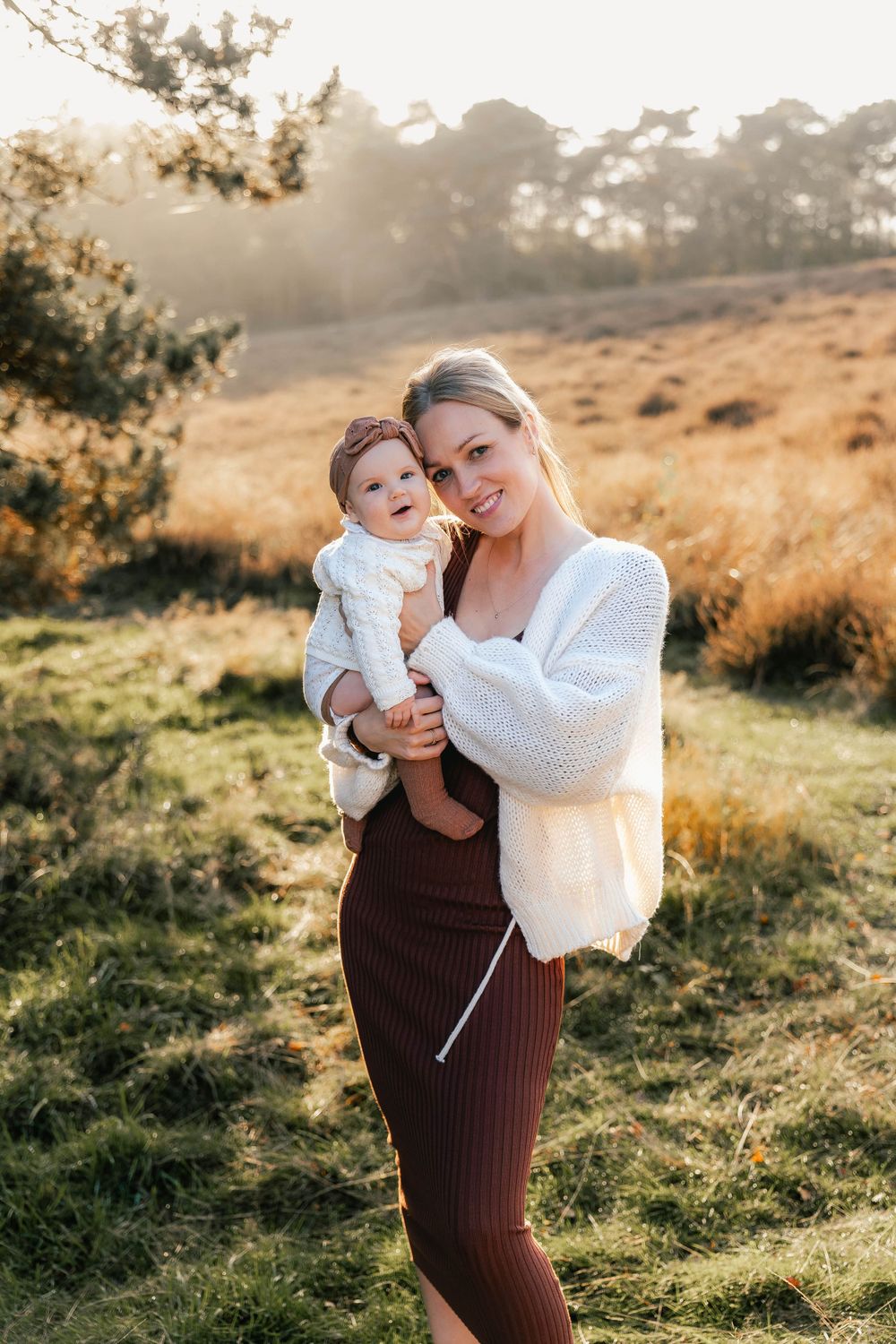 A candid outdoor portrait of parent and baby wearing white on a grassy hillside at sunset.