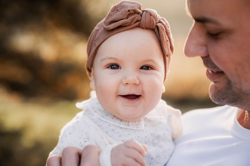 Close up of smiling baby in white dress and pink headband with adult.
