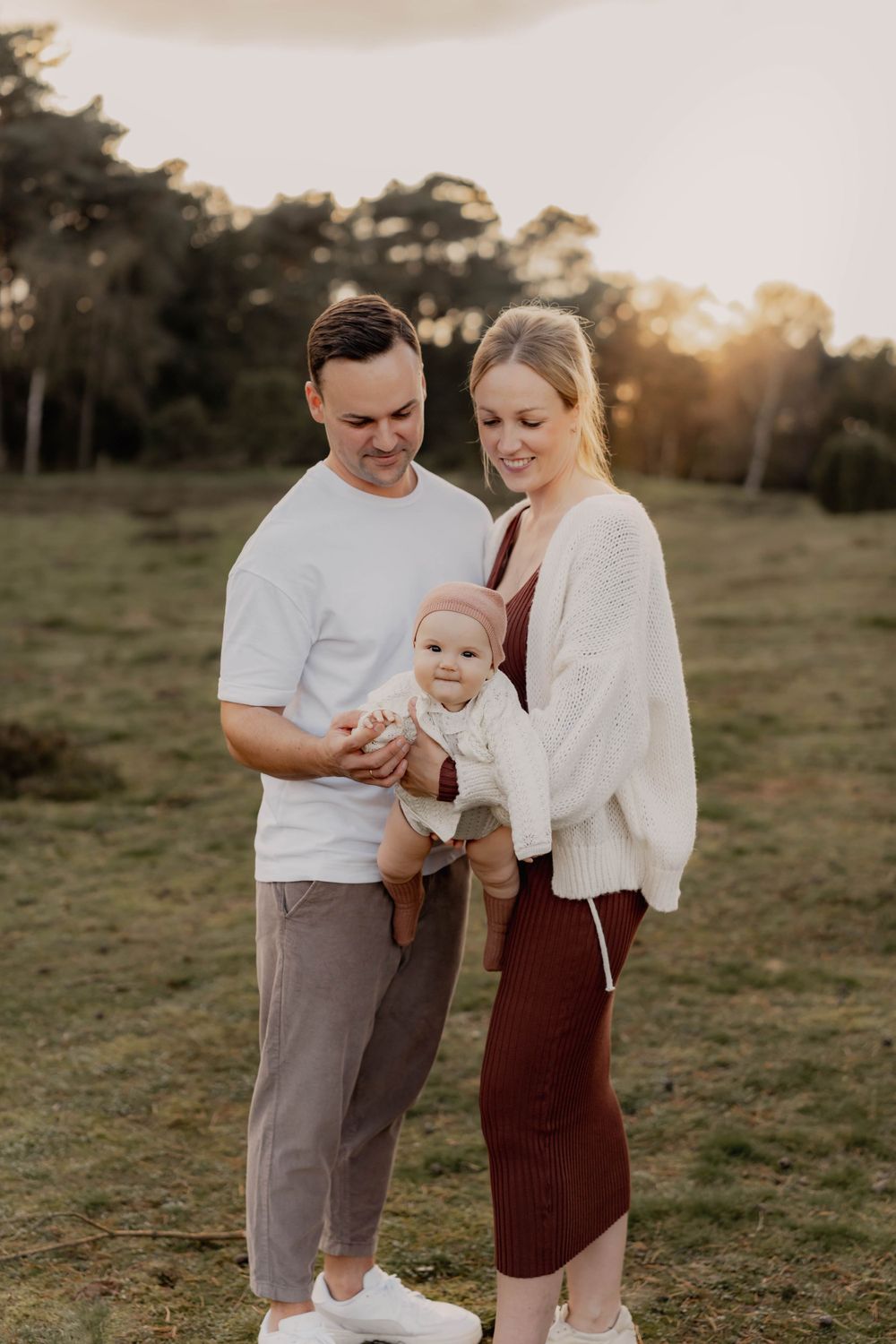 Intimate family portrait outdoors with baby in white against a golden sunset backdrop.