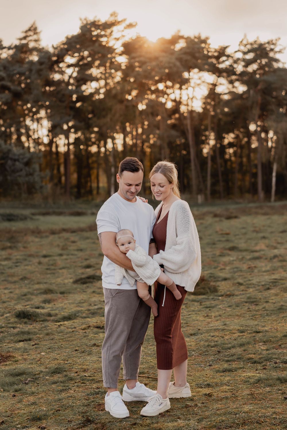 A full-length family portrait standing together in a field with pine trees at golden hour.