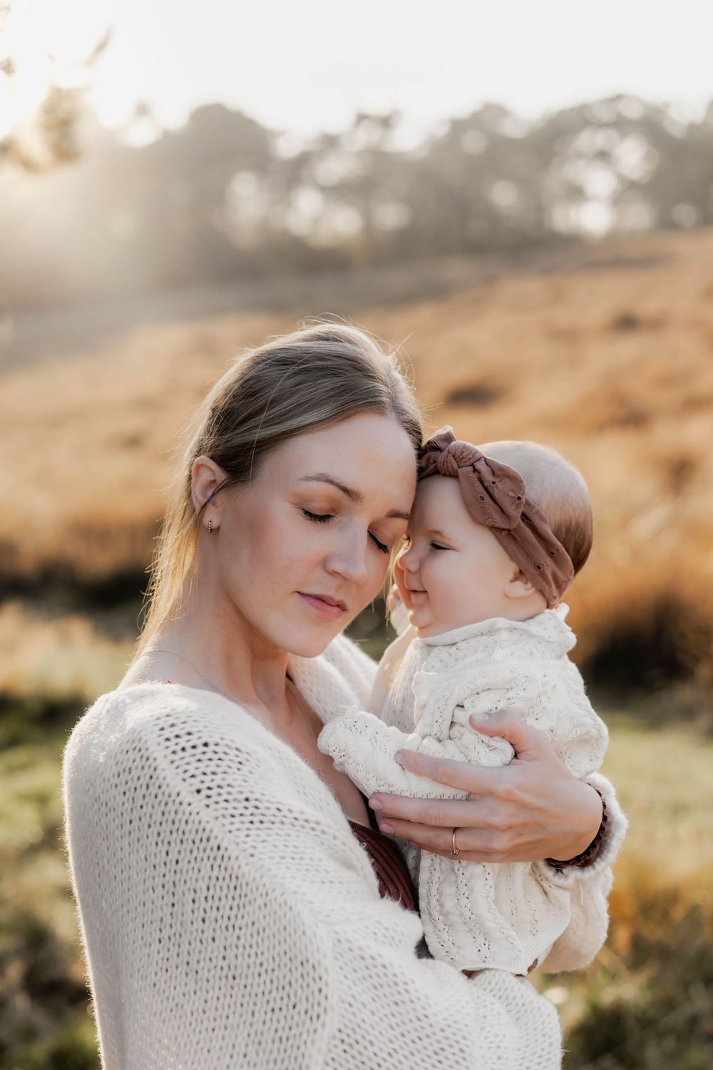 Intimate moment between parent and baby in white sweaters during golden hour.