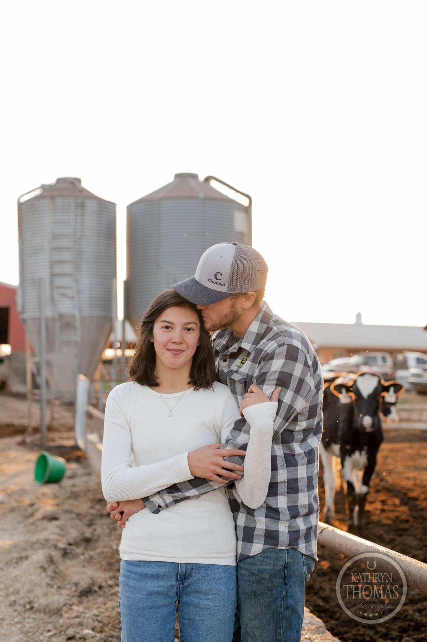 A sweet moment shared between two people on a farm with grain silos and dairy cows behind them.