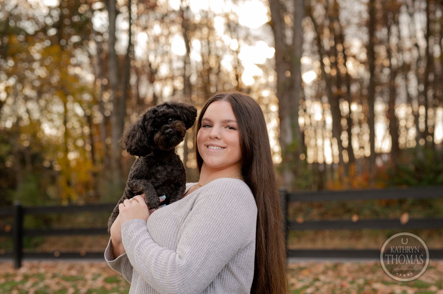 Someone cuddles a small black dog while wearing a light sweater in an autumn setting.