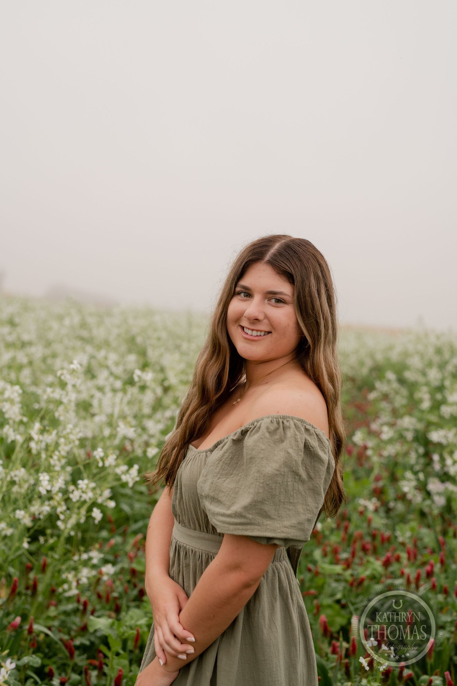 Model wearing an olive green dress stands in a field of white and red flowers on an overcast day.