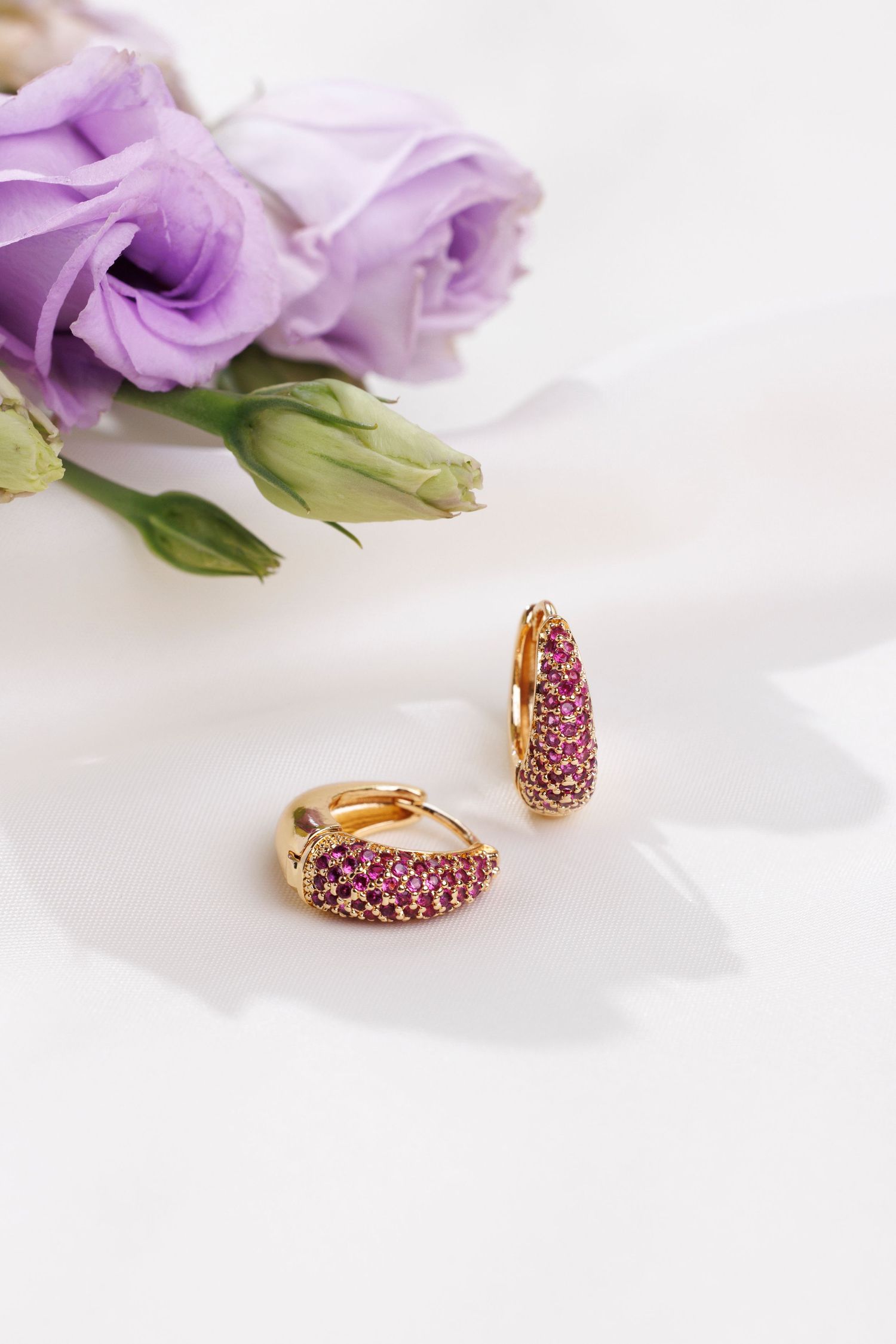 Purple rose flowers with gold and pink crystal-encrusted hoop earrings on white satin backdrop.