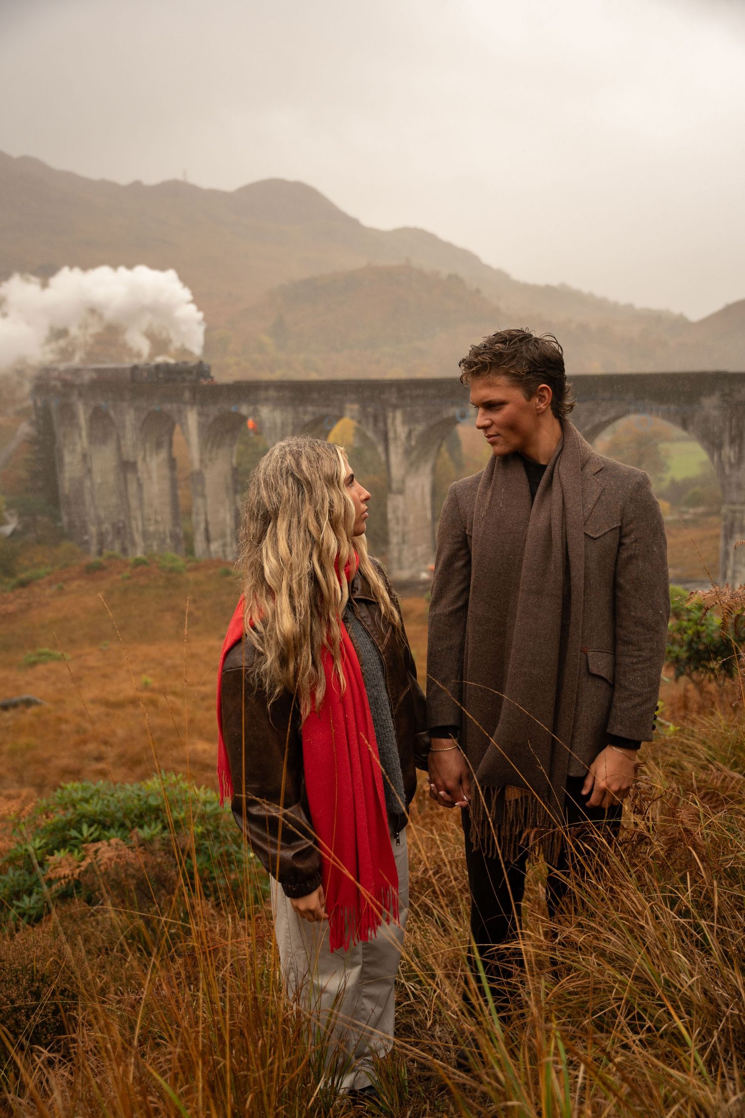 Cinematic shot of a couple in front of the Harry Potter train filming location in Fort William, Scotland, with dramatic, magical vibes.
